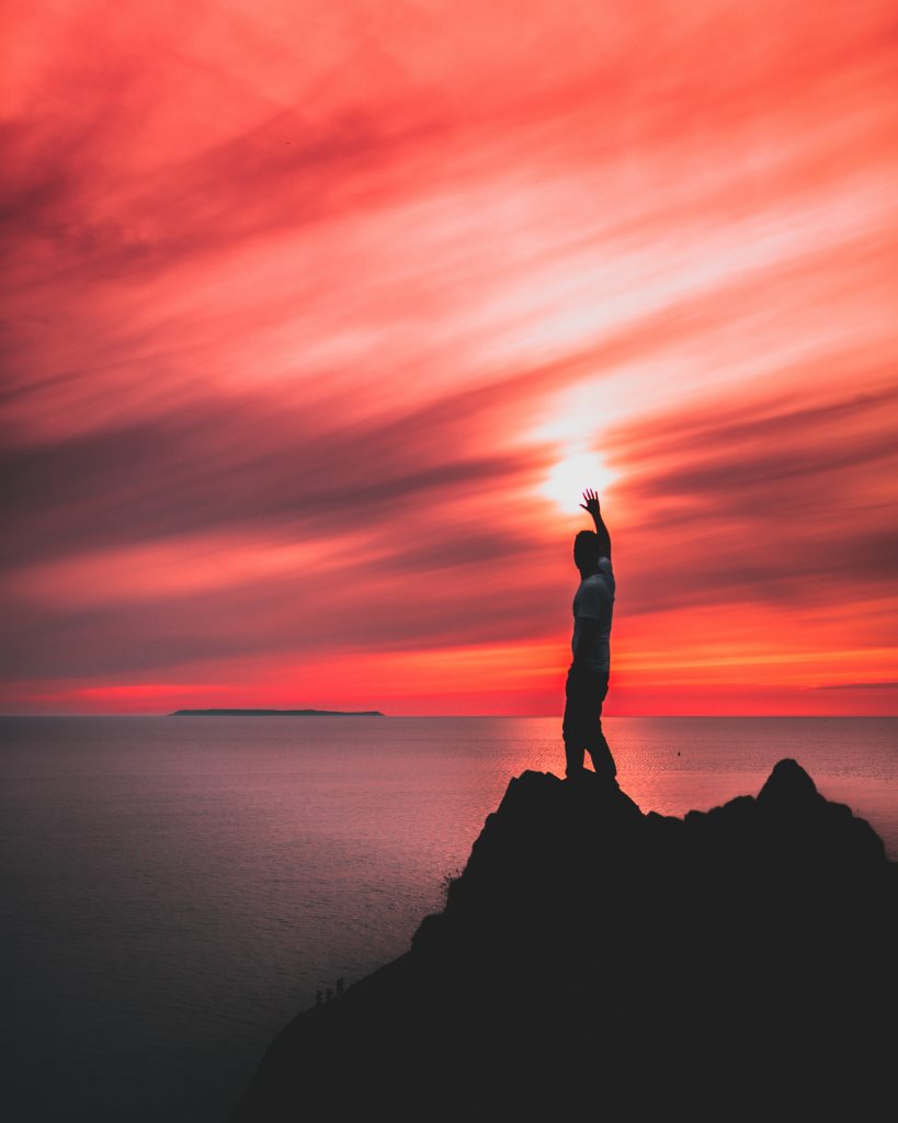 A person standing on a rock beside a sea, holding his right hand up towards the sun shining in a red sky - AAAF