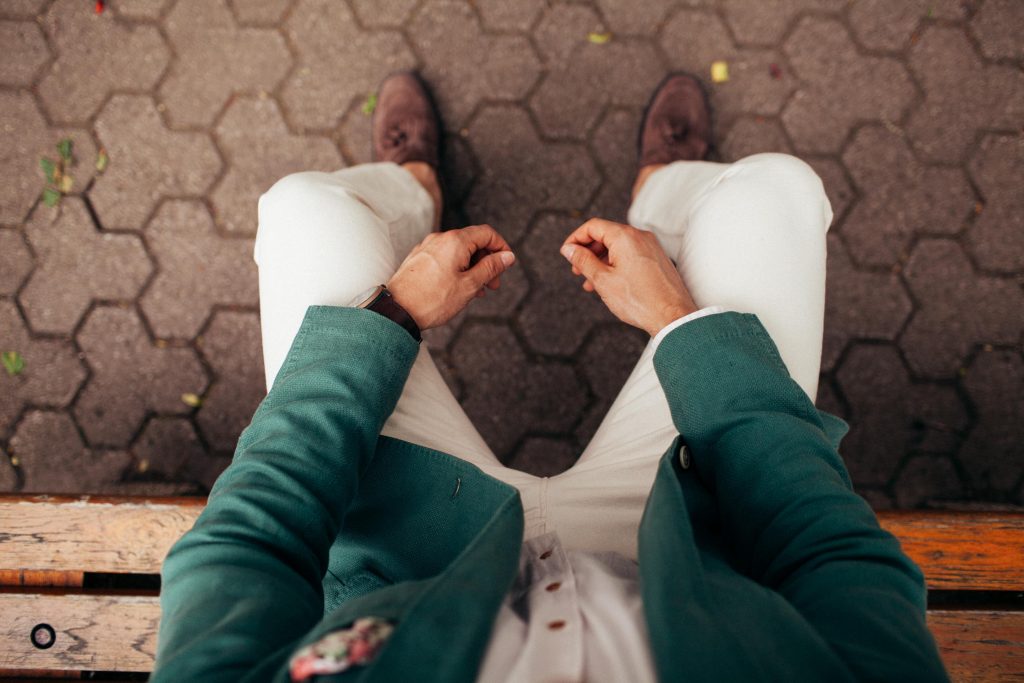 Top view of a man sitting on a bench wearing green coat & white pant, with brown shoes, resting his hands on legs - AAAF
