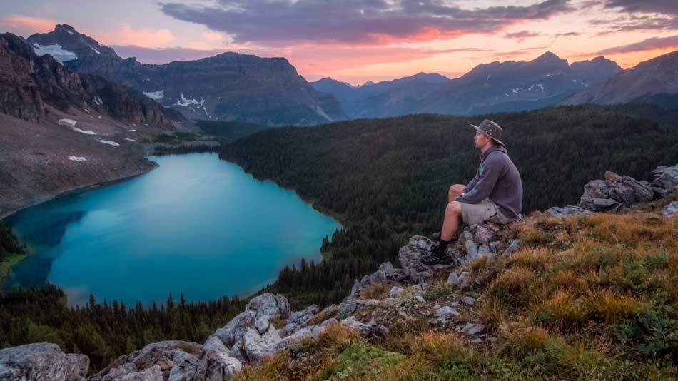A man sitting on rocks near a lake next to a forest, looking at the mountain range across - AAAF