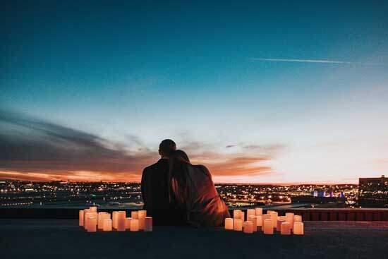 Husband & Wife in love, sitting together with candles around them, looking at the wide expanse of city lights & blue sky