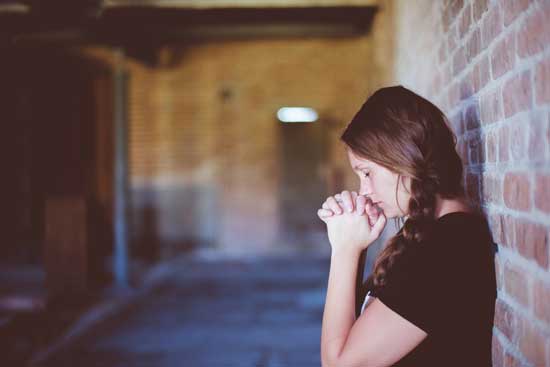 A woman closes her eyes and raises her hands in prayer as she leans her back against a brick wall.