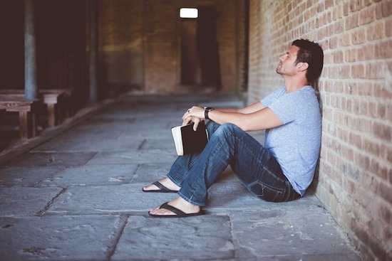 A man leaning against a wall, holding a Bible, and praying to Jesus Christ in faith