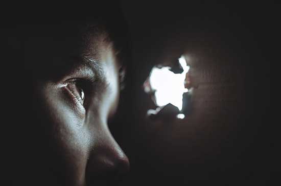 A person in the dark peaking through a hole that's letting in light