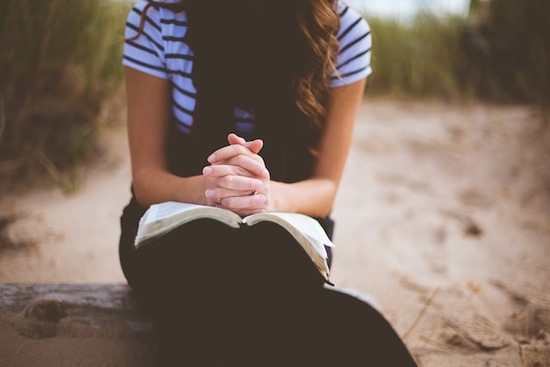 A modestly dressed woman with her hands folded in prayer over her open Bible.