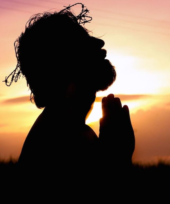 An orange sunset rises behind a silhouette of Jesus as He clasps His hands in prayer and wears a crown of thorns.