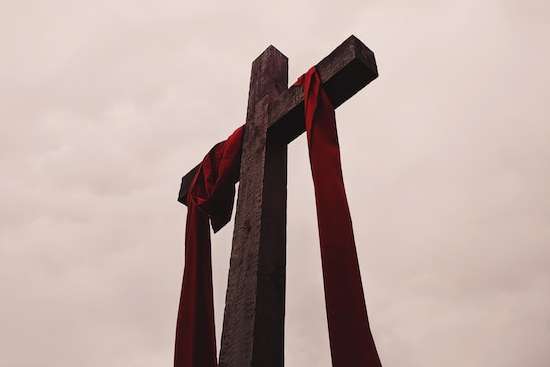 A wooden cross draped with red cloth stands before a cloudy sky.