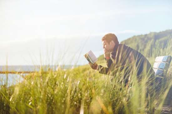 A man leaning forward and reading the Bible as he sits in a grassy field overlooking a lake