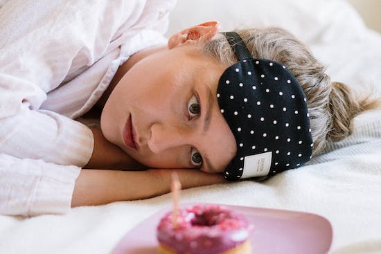 A girl laying in bed and resisting the temptation to eat a donut on a plate in front of her