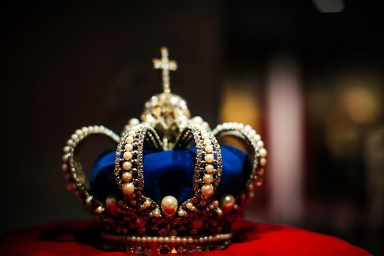 A blue crown, studded with pearls and topped with an emblem of a cross, sits on a red pillow.