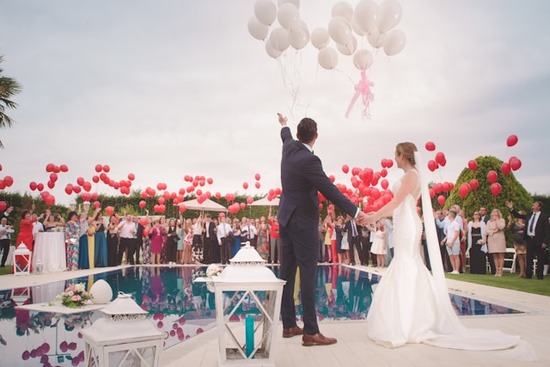 A bride and groom release white balloons into the air at their wedding celebration as all their guests cheer.