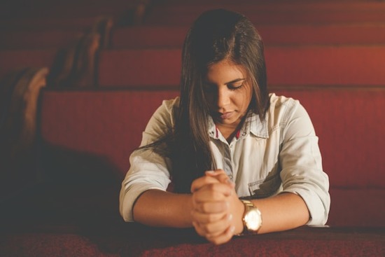 A girl sitting in a church pew, folding her hands and closing her eyes in prayer.